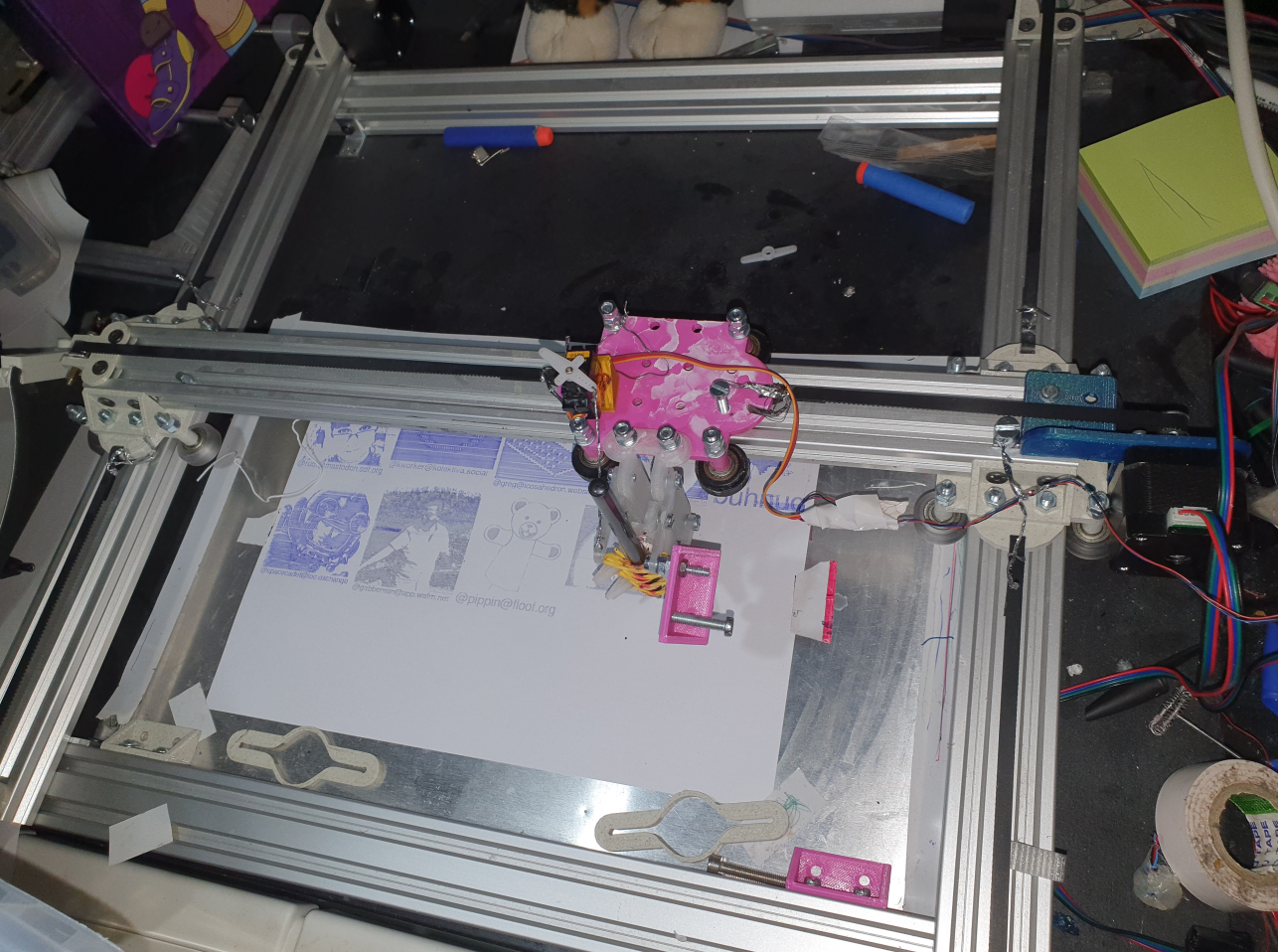 A pen plotter consisting of aluminum extrusions and 3D printed wheels with 2 Y motors and one X motor and a servo lifting and putting down a pen and an A4 pages is being plotted on below it